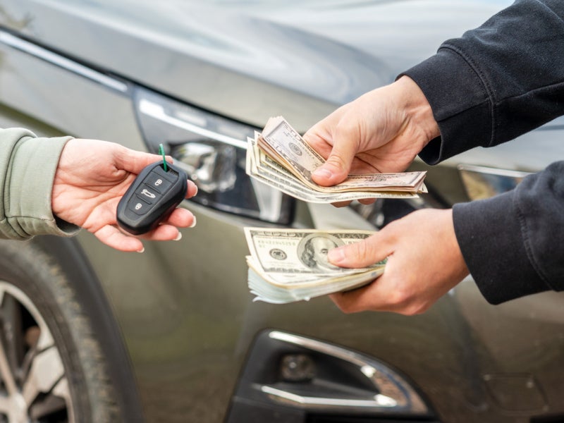 Two people exchange car keys and cash in front of a vehicle, suggesting a car sale. Hands are visible, conveying a transactional moment.